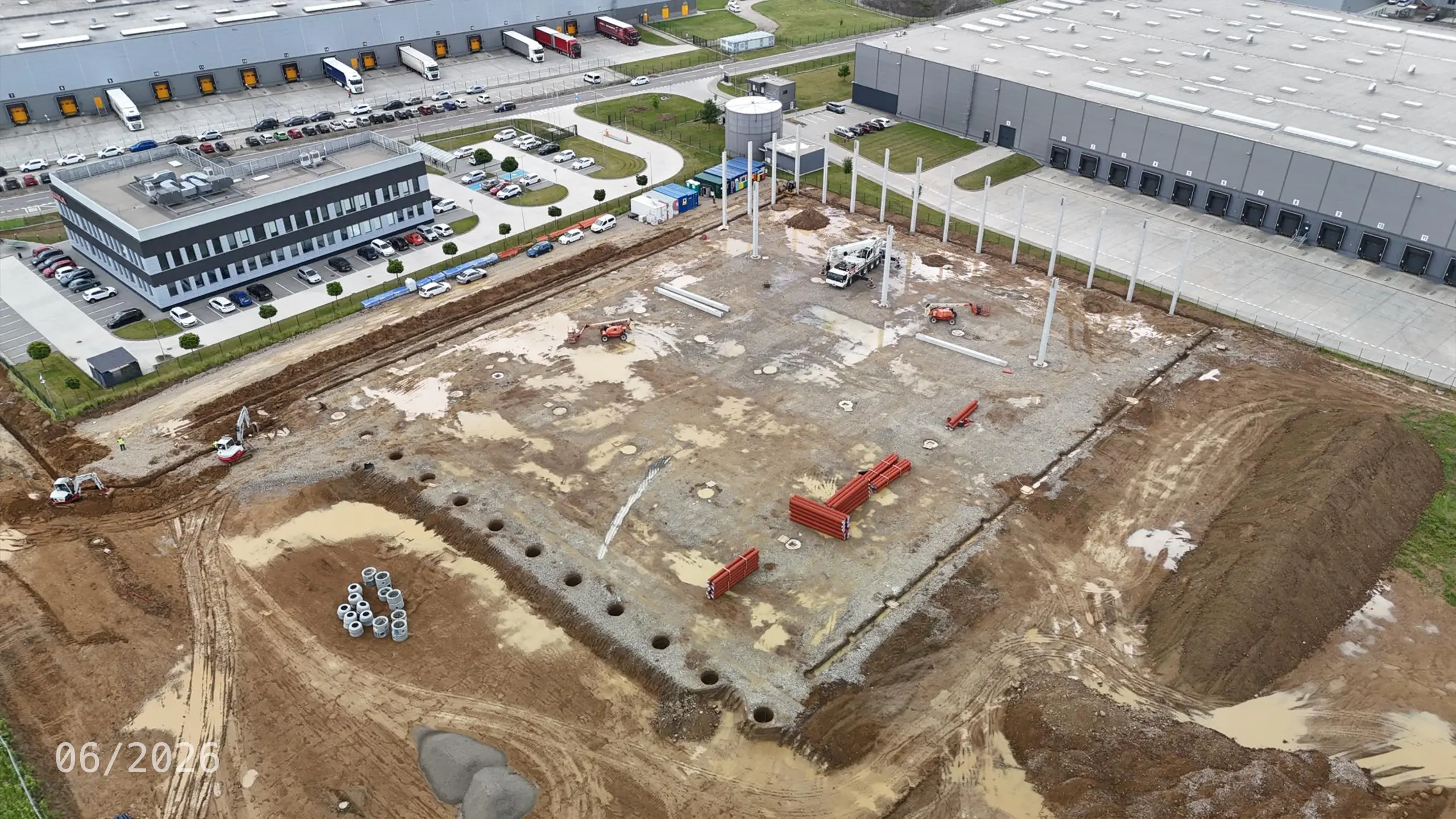 Priemyselný projekt Dubnica 4 – výstavba nosnej konštrukcie novej skladovej a administratívnej haly v Dubnici nad Váhom. Na fotografii sú viditeľné realizované základy a montáž železobetónových stĺpov, ktoré tvoria základ budúceho logistického objektu nadväzujúceho na existujúce budovy priemyselného parku. - obrázok 9