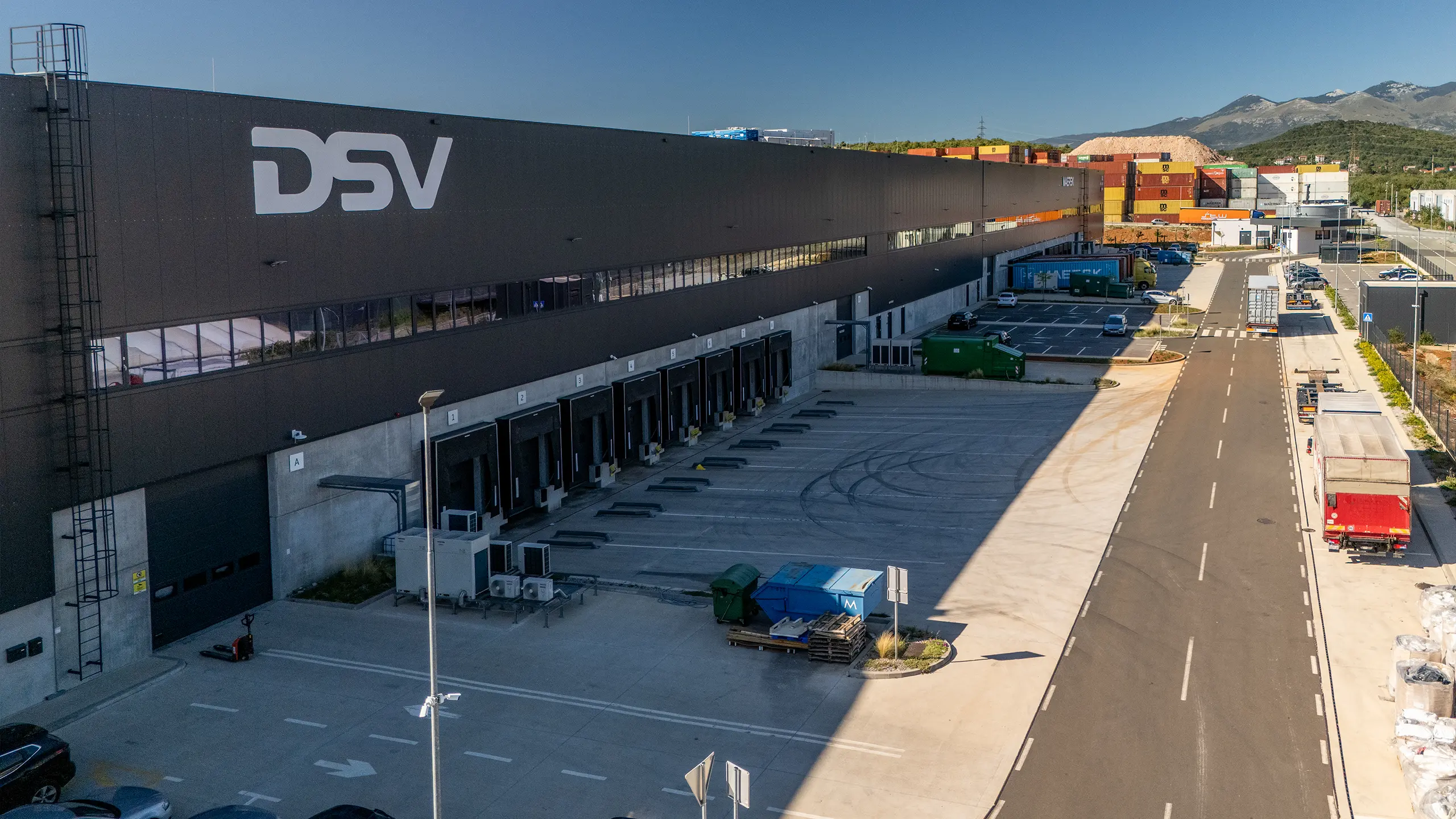Logistics hall Rijeka 1 - a view of a modern hall from a bird's eye view with dark gray cladding, roof photovoltaic panels, large parking lot, logos on the facade: DSV, MAERSK, Logexpert and Sympathy. - obrázok 2