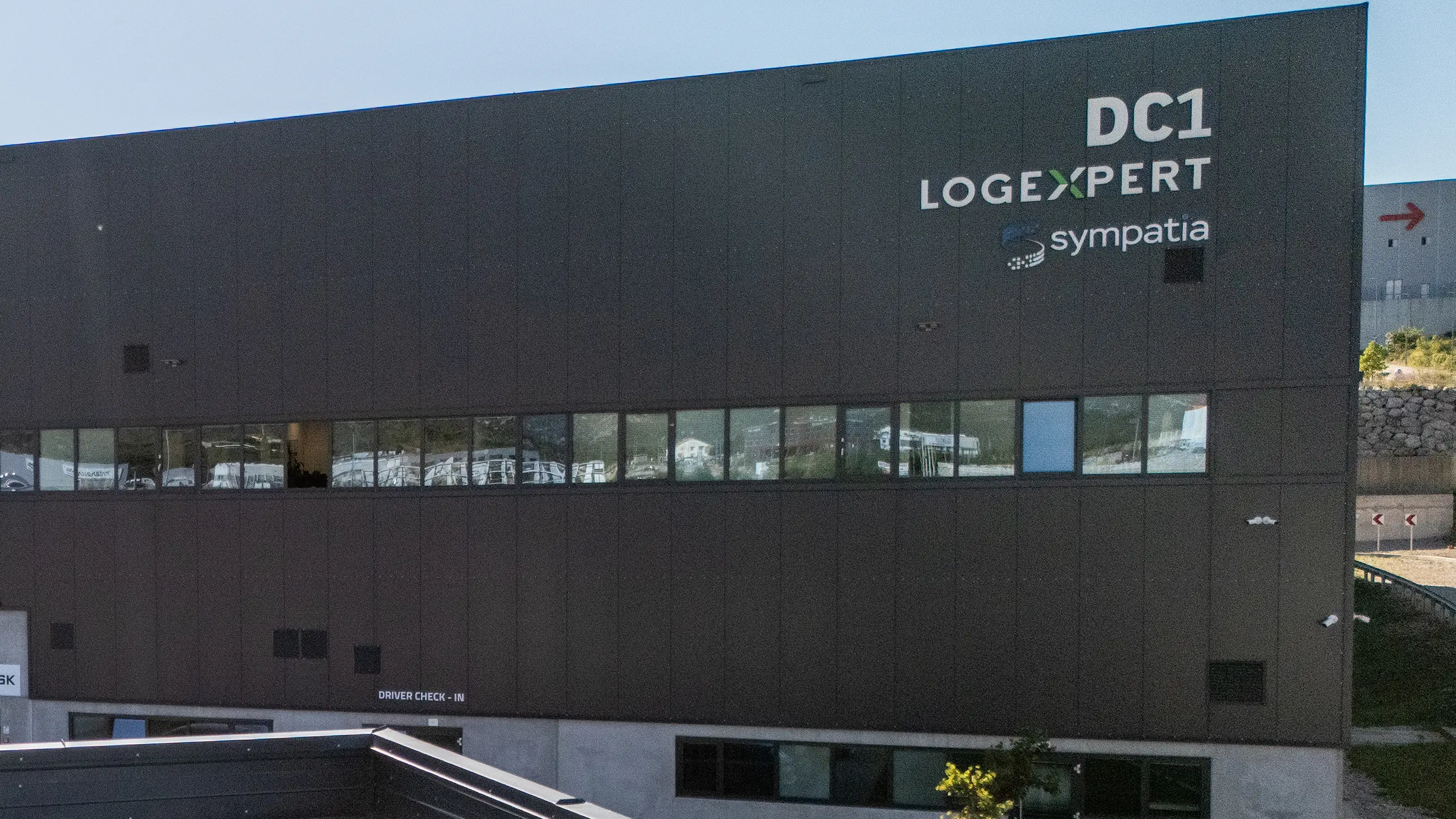 Logistics hall Rijeka 1 - a view of a modern hall from a bird's eye view with dark gray cladding, roof photovoltaic panels, large parking lot, logos on the facade: DSV, MAERSK, Logexpert and Sympathy. - obrázok 3
