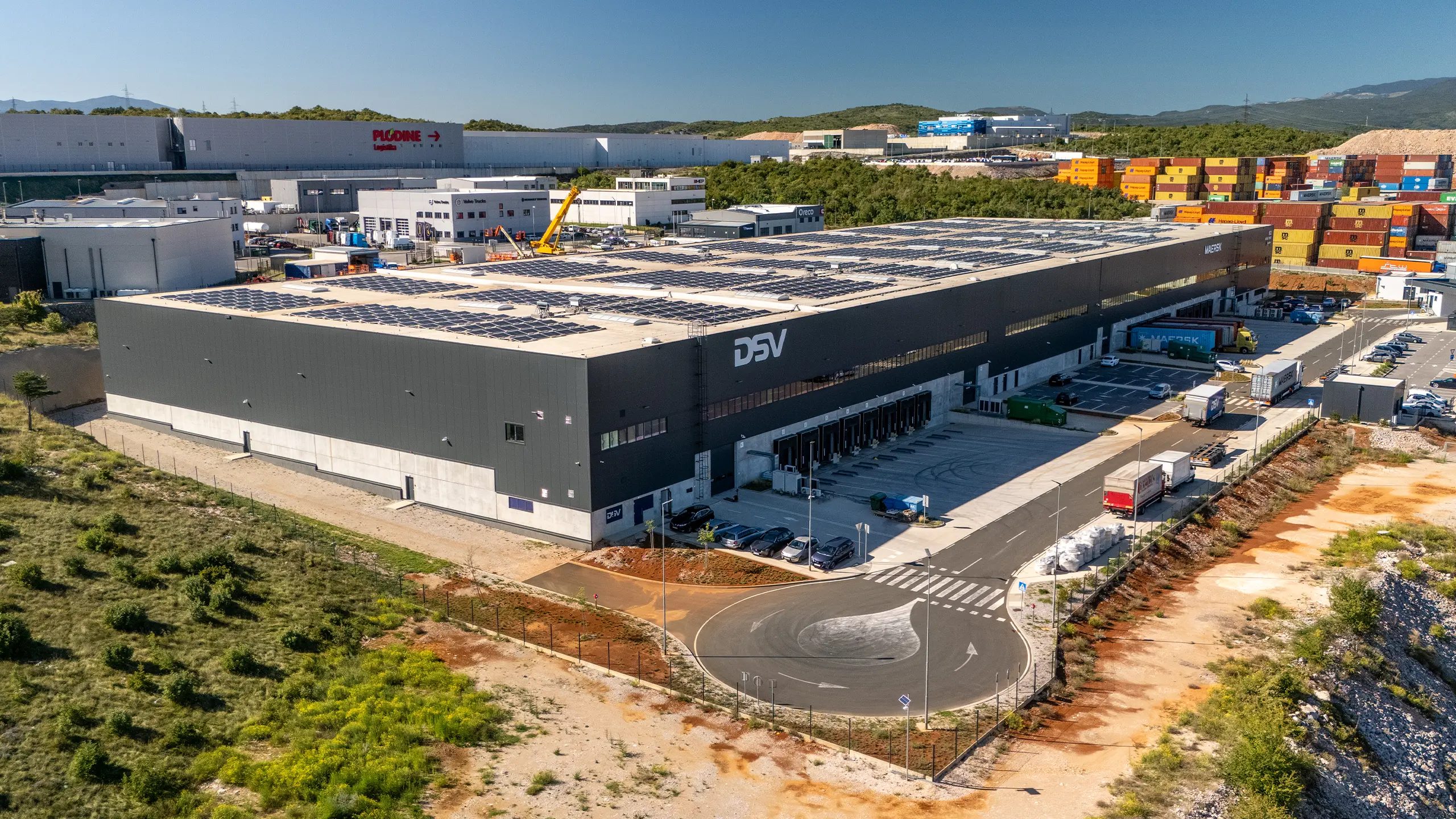 Logistics hall Rijeka 1 - a view of a modern hall from a bird's eye view with dark gray cladding, roof photovoltaic panels, large parking lot, logos on the facade: DSV, MAERSK, Logexpert and Sympathy. - obrázok 4