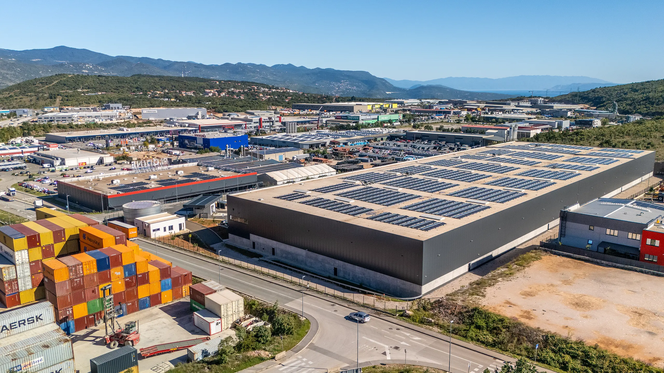 Logistics hall Rijeka 1 - a view of a modern hall from a bird's eye view with dark gray cladding, roof photovoltaic panels, large parking lot, logos on the facade: DSV, MAERSK, Logexpert and Sympathy. - obrázok 5