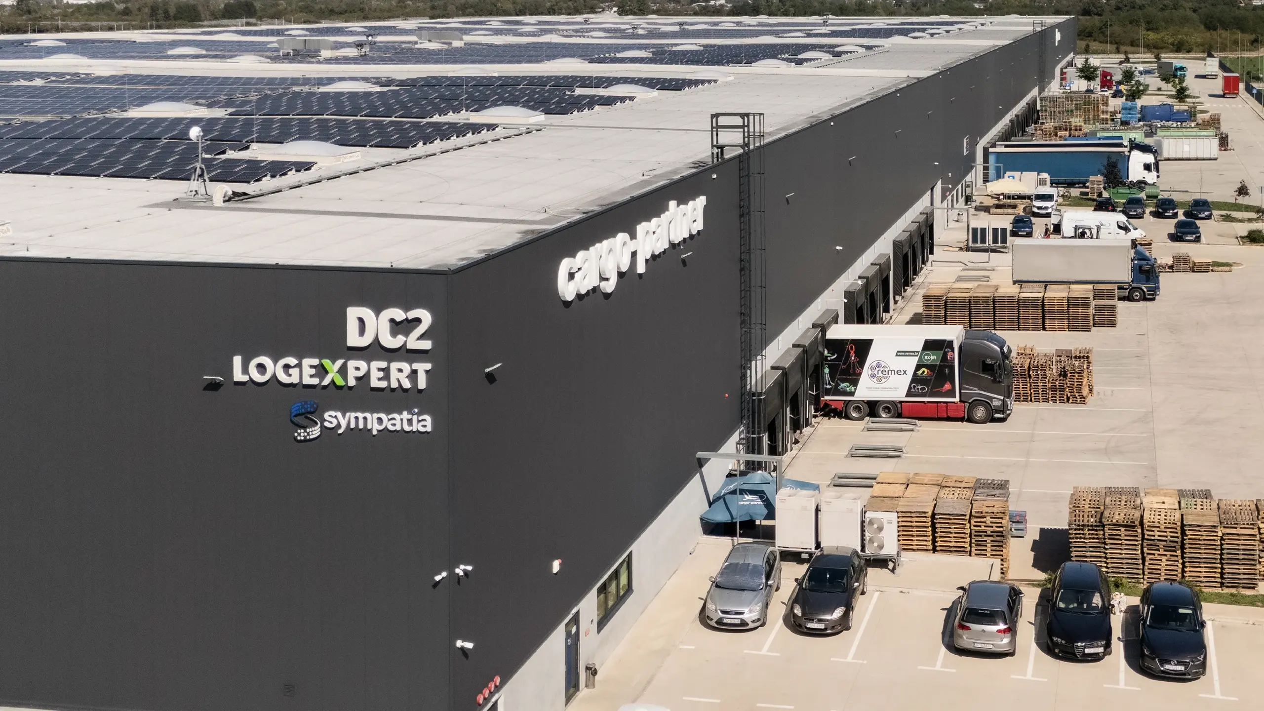 Priemyselný projekt Záhreb 1 – moderný logistický park pri Záhrebe s vysokokapacitnými skladovými a distribučnými priestormi. Letecký pohľad zachytáva rozsiahlu logistickú halu s nakladacími rampami, manipulačnými plochami pre nákladnú dopravu a fotovoltickými panelmi na streche, navrhnutú podľa najvyšších priemyselných štandardov pre logistické, retailové a automotive spoločnosti. - obrázok 2