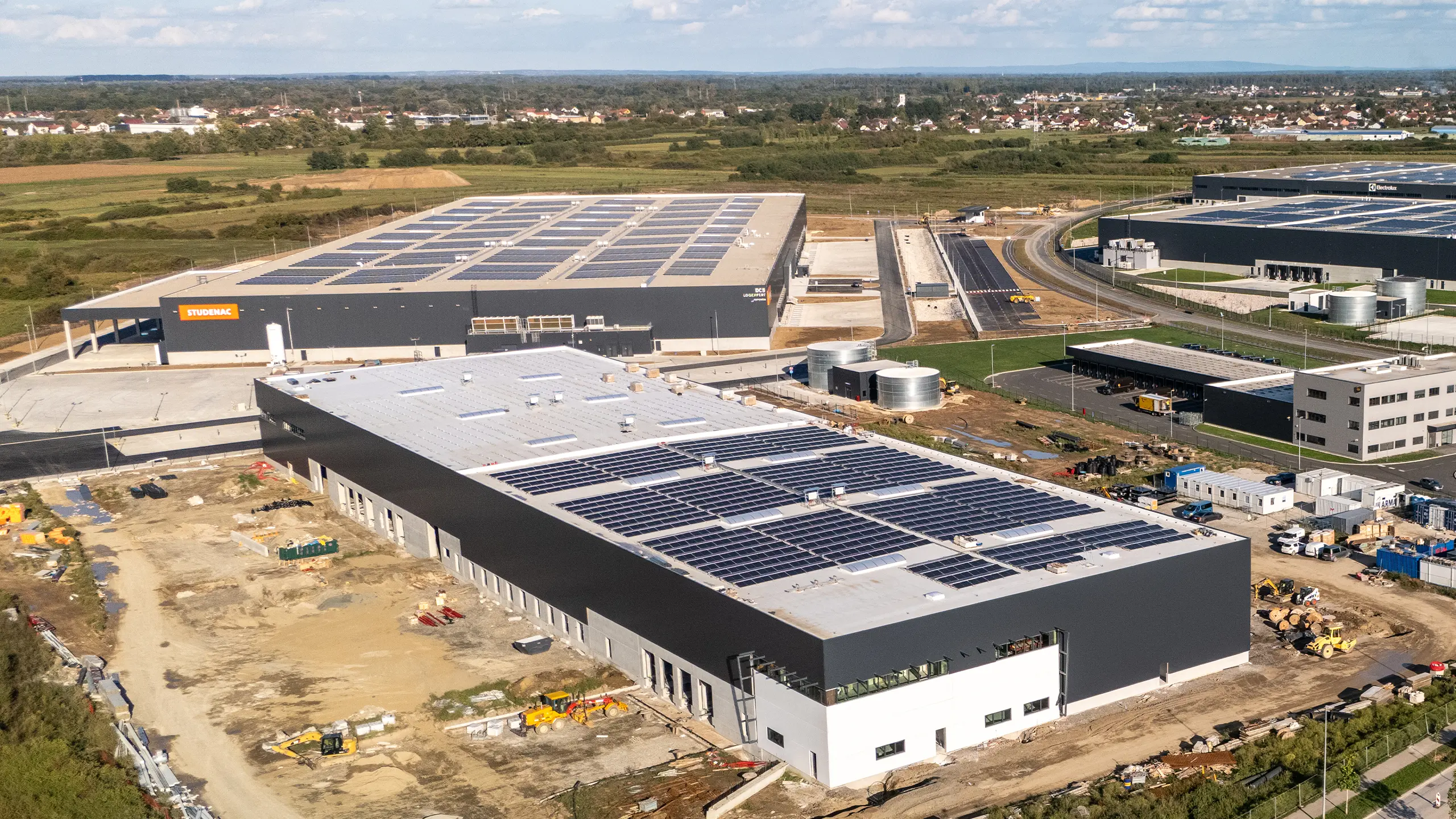 Industrial Project Zagreb 3 – aerial view of the expanding third phase of the Zagreb logistics park. In the foreground is a newly built logistics facility nearing completion with rooftop photovoltaic panels, while a completed warehouse bearing the Studenac logo is visible in the background. The project provides high-standard warehouse space, including refrigerated and deep-freeze facilities, with a strong focus on energy efficiency and sustainable solutions. - obrázok 3