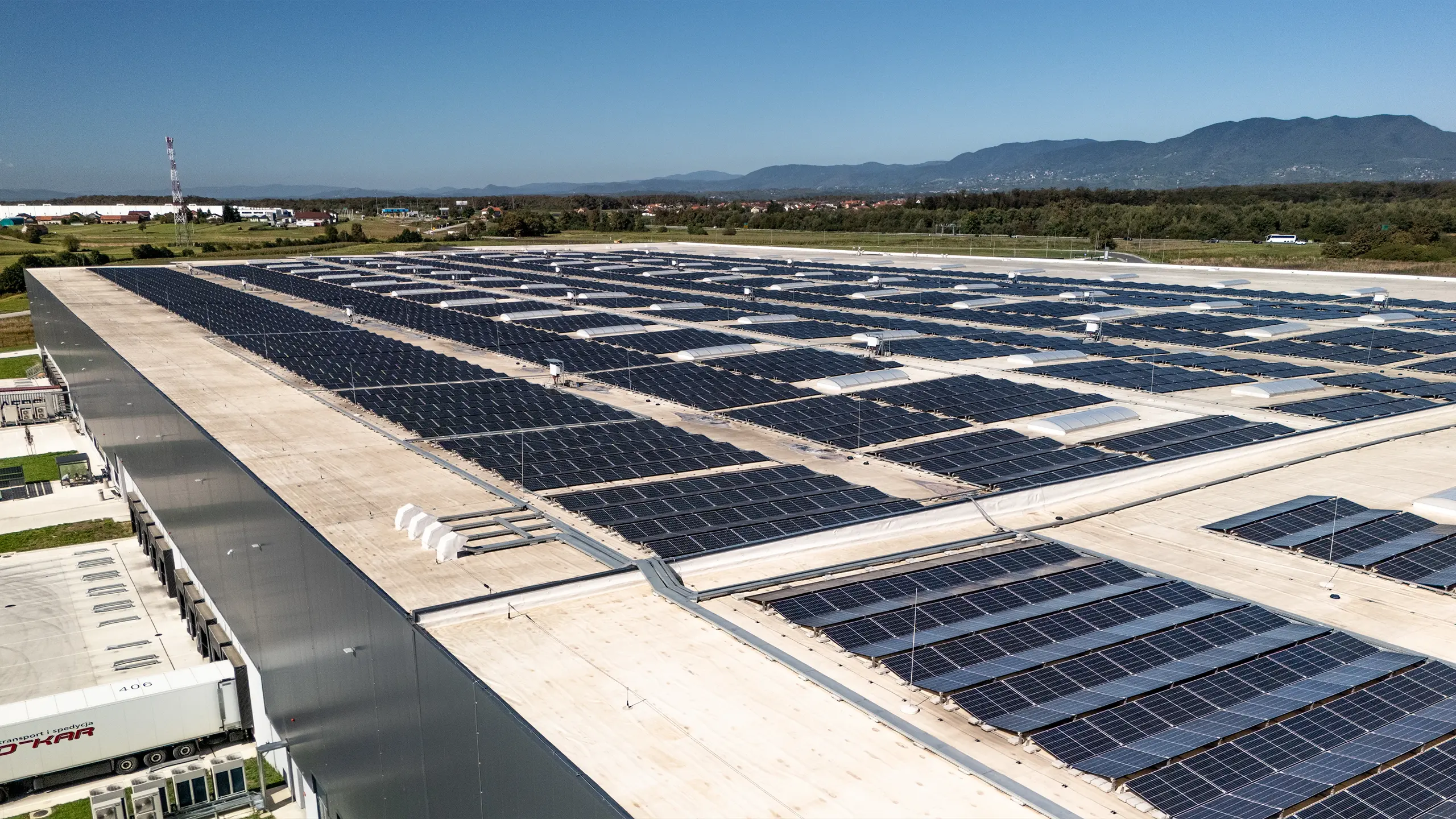 Industrial project Zdenčina 1 – detailed view of the rooftop photovoltaic power plant installed on a logistics facility within the regional distribution centre. The extensive solar panel system supports the project's energy efficiency and fulfils the environmental standards of modern industrial real estate. The surrounding landscape and mountain range near Zagreb can be seen in the background. - obrázok 4