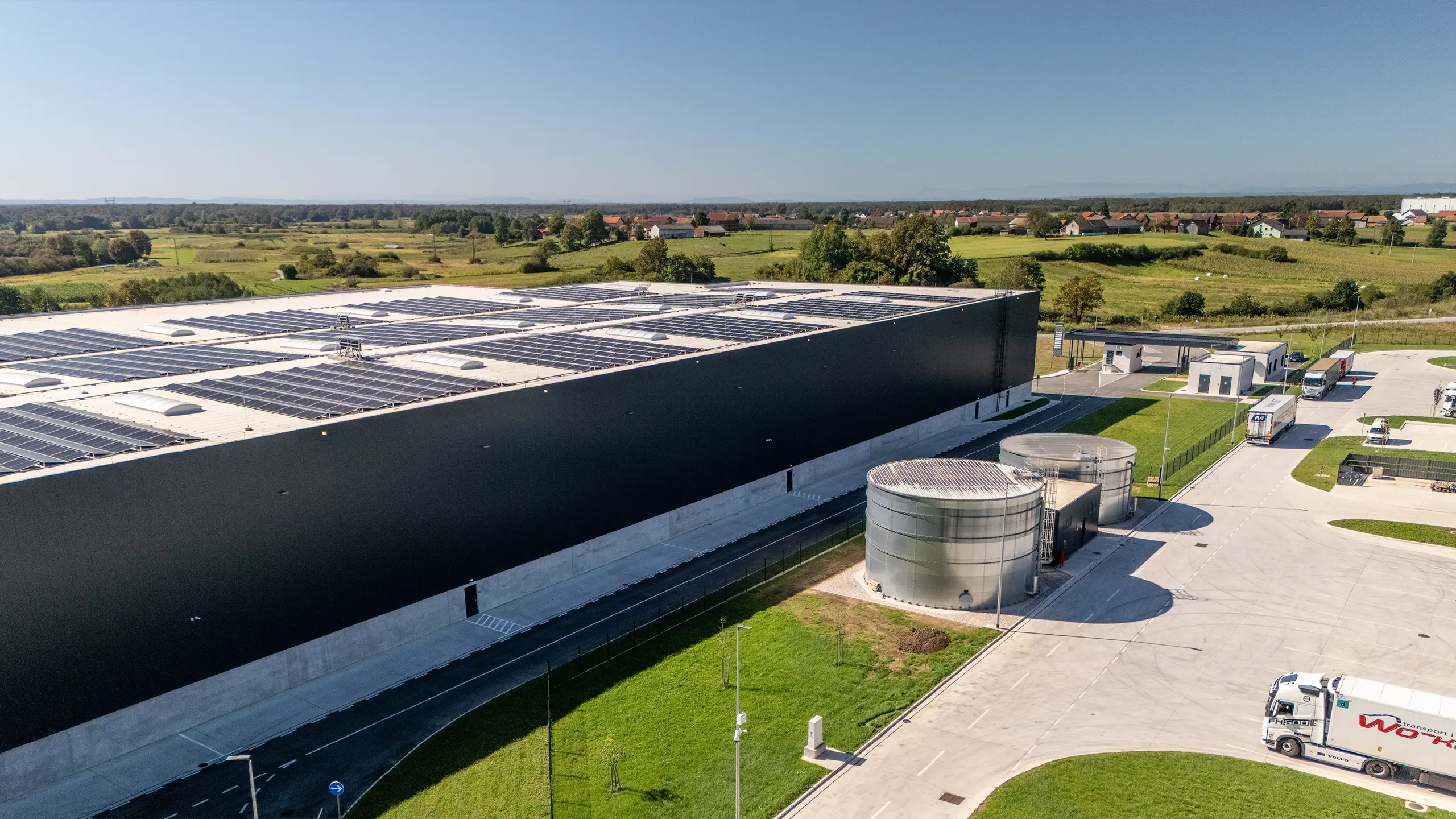 Industrial project Zdenčina 1 – view of a logistics facility within the regional distribution centre featuring rooftop photovoltaic panels and supporting site infrastructure. The project includes retention tanks, modern transport solutions and extensive operational areas that support efficient warehouse operations. Developed according to sustainable construction principles, the facility meets high environmental and logistics standards. - obrázok 5