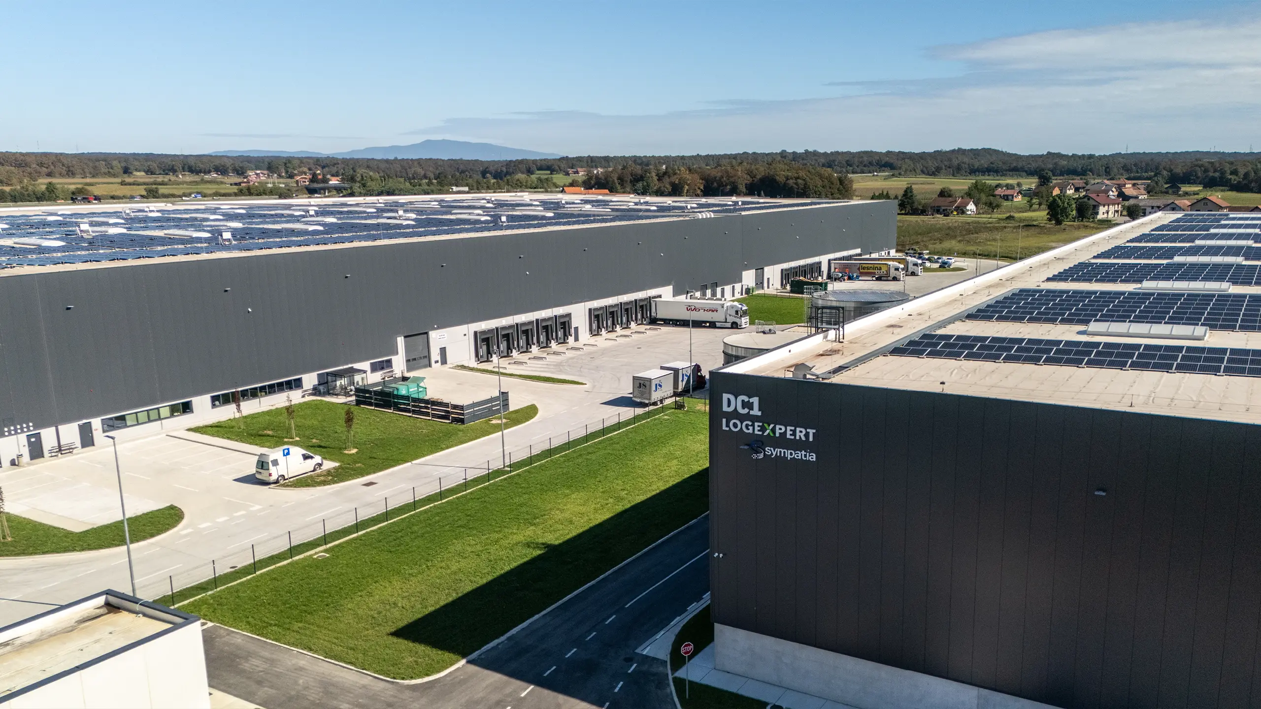 Industrial project Zdenčina 1 – view of a logistics facility within the regional distribution centre featuring rooftop photovoltaic panels and supporting site infrastructure. The project includes retention tanks, modern transport solutions and extensive operational areas that support efficient warehouse operations. Developed according to sustainable construction principles, the facility meets high environmental and logistics standards. - obrázok 7